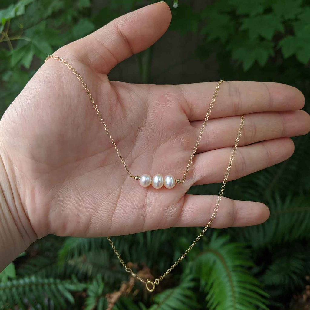 A necklace with 3 creamy coloured pearls on a dainty gold chain resting on a hand for scale