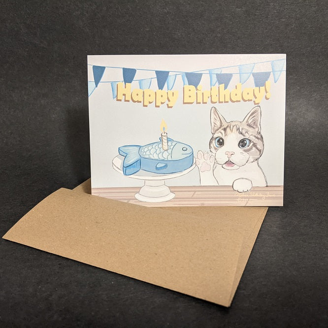 This greeting card shows a grey and white cat reaching out to his fish shaped birthday cake